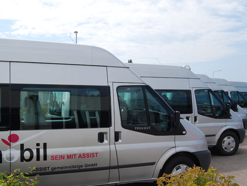 5 ASSIST-Firmenbusse parken in einer Reihe mit dem ASSIST-Logo und der Aufschrift "Mobil sein mit ASSIST" an der Bus-Seite.